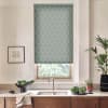 Patterned teal roller blind covers window above stainless sink, filtering light; kitchen countertop holds bowl of leafy greens, folded towel, potted plants, framed sketch on pale wall, wooden cabinets below.