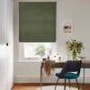 A green Roman blind, fully lowered with slight folds, covering a sash window and filtering soft daylight; desk beneath with vase of flowers and teal-upholstered chair in bright study.