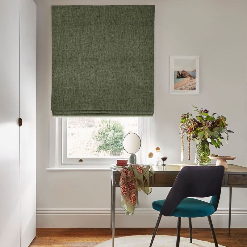 A green Roman blind, fully lowered with slight folds, covering a sash window and filtering soft daylight; desk beneath with vase of flowers and teal-upholstered chair in bright study.