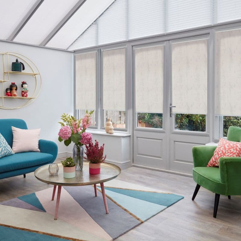 Light-beige patterned roller blinds across tall glass doors and upper triangular windows, partially lowered and filtering daylight; modern sunroom with teal sofa, green armchair, round coffee table and floral arrangement.
