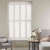 White plantation shutters covering a tall recessed window—four narrow hinged panels with horizontal louvers—closed, filtering soft daylight; neutral bedroom setting with upholstered armchair, bedside table, lamp and headboard.