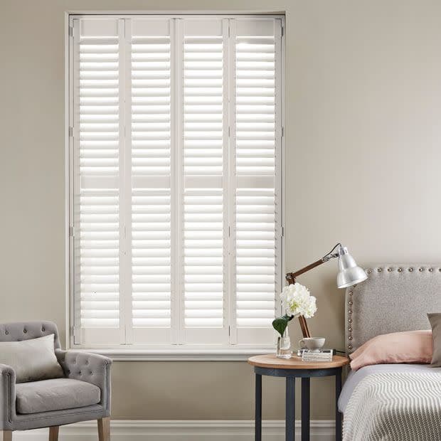 White plantation shutters covering a tall recessed window—four narrow hinged panels with horizontal louvers—closed, filtering soft daylight; neutral bedroom setting with upholstered armchair, bedside table, lamp and headboard.