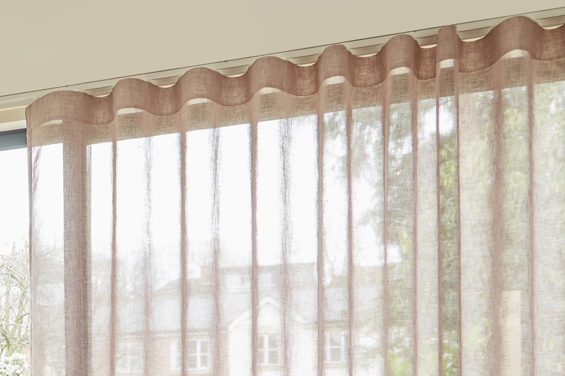 Sheer beige wave-headed curtains (ripple-fold) — hanging full-length, forming soft vertical pleats — filtering bright daylight into an interior living space, partially revealing blurred houses and trees outside.