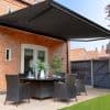 Large black retractable awning mounted above patio doors, fully extended with metal arms and LED trim; shading a wicker dining set on a brick-walled backyard patio under clear daylight.