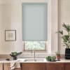 Window with pale blue roller blind covers most pane, revealing greenery below; set above a kitchen sink and faucet, with wooden cabinets, potted plants and a framed sketch.