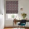 A geometric-patterned Roman blind (burgundy, grey, white) lowered over a window, filtering daylight above a desk with teal chair, vase of flowers and framed seaside print in a bright office.