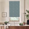 Blue floral roller blind hangs down, filtering daylight through a kitchen window; stainless faucet below, wooden cabinets and countertop with bowls of greens, potted plant and framed sketch.