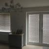 Horizontal venetian blinds on a window and patio door, closed and filtering dim daylight; living-room corner with a small cabinet, owl ornament, two photos, and chandelier.
