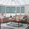 Teal cellular blinds covering tall conservatory windows, fully lowered pleated honeycomb panels filtering soft daylight; setting a bright conservatory living room with beige sofa, wooden coffee table, and textured rug.