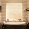 Cream horizontal Venetian blinds covering a bathroom window, fully lowered and closed, diffusing warm daylight onto an oval bathtub by striped beige walls with shelves, towels and decorative items.