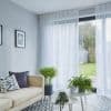 Sheer white floor-to-ceiling curtains on a ceiling track, filtering daylight through sliding glass doors; bright living room with beige sofa, patterned tile floor, plants, garden and shed view.