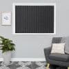 Vertical dark grey blinds cover a white-framed window, fully closed with beaded chain weights; pale grey living-room wall flanked by a potted plant left and an upholstered armchair right.