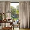 Floor-to-ceiling pleated patterned beige curtains, parted centrally to reveal a glass door; curtains hang in folds, filtering daylight into a dining area with wooden table, chairs, pendant lamp and garden view.