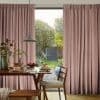 Pleated dusty-pink floor-length curtains with subtle texture, parted at the center to reveal a sunlit garden; a wooden dining table set for breakfast, pendant lamp and woven chairs.