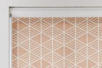 A closed roller blind with a peach-and-white geometric diamond pattern, blocking light, mounted in a white cassette with a bead chain at left against a brightly lit interior wall.