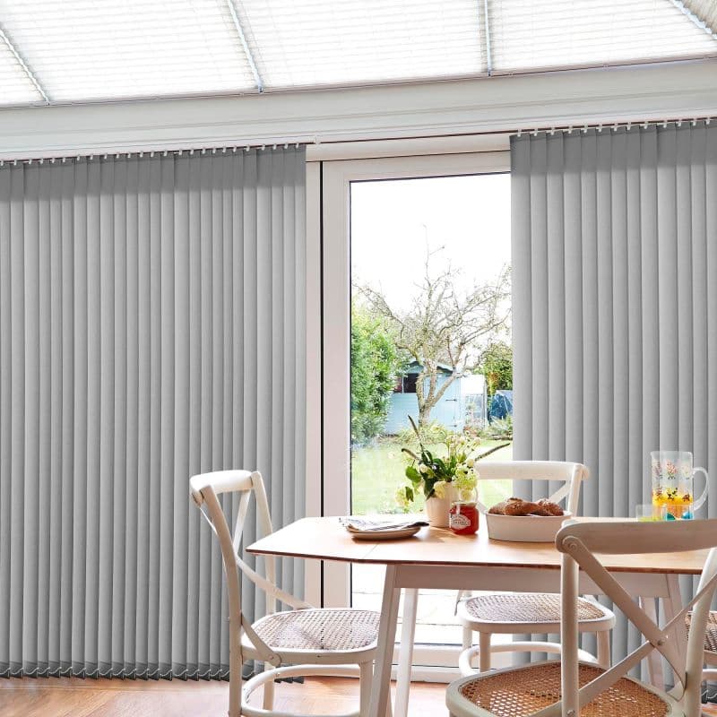 Grey vertical blinds (floor-to-ceiling) flank sliding glass doors, partially open and filtering daylight; a sunlit dining area with wooden table, chairs, vase of flowers, and a green garden outside.
