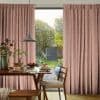 Pink pleated full-length patterned curtains, drawn to each side revealing a central glass patio door; filtering soft daylight into a dining area with wooden table, pendant lamp and garden.