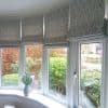 Leaf-patterned fabric Roman blinds across a bay of windows, partially lowered with visible pull chains, softly filtering daylight; sill holds a jug of flowers, view of manicured shrubs outside.