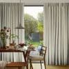 Floor-to-ceiling beige pleated curtains, parted centrally to expose glass door, softly filtering daylight; framing a dining table with pendant light, plates of pancakes, a chair, and a sunlit garden view.