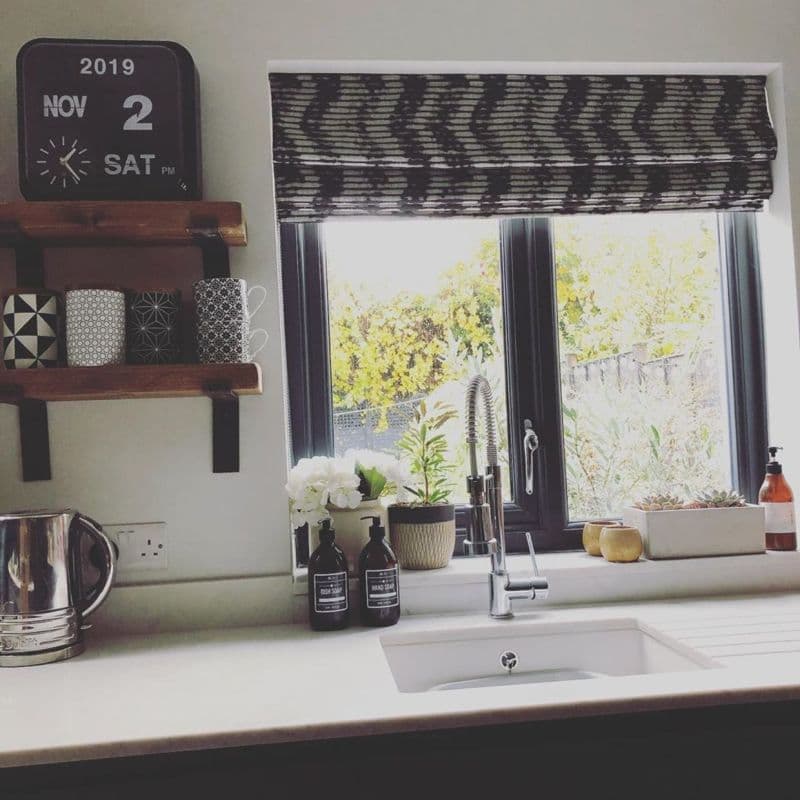 Patterned fabric Roman blind, partially lowered across a black-framed double window, filtering bright daylight; stainless spring-tap over kitchen sink, potted plants, soap bottles and wooden mug shelves nearby.
2019
NOV
2
SAT
PM
DISH SOAP
HAND SOAP