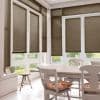 Brown horizontal Venetian blinds covering bay windows and glass doors, lowered and partially filtering daylight; bright dining nook with white table and chairs, floral cushions, tiled floor and garden view.