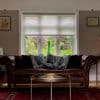 Translucent white roller blind, lowered halfway and filtering daylight; in a living room behind a brown leather sofa with patterned cushions; view of lush garden and stone wall.