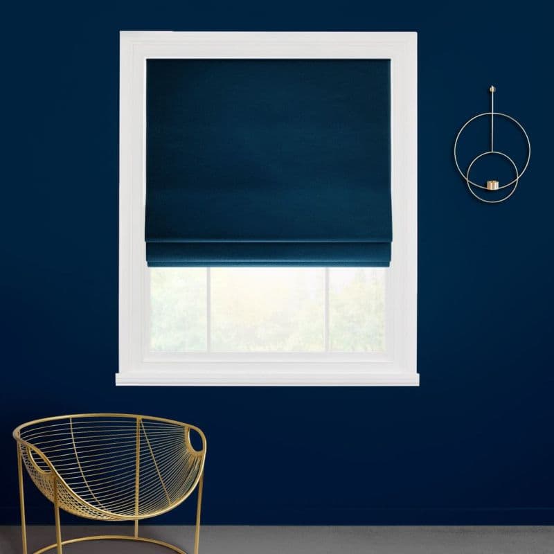 Navy Roman shade, partially lowered over a white-framed window, filtering daylight; set in a dark-blue wall with a gold chair below and a circular gold wall sconce to the right.