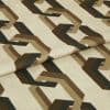 Cream fabric sample printed with repeating brown and black interlocking geometric shapes; folded across the center; close-up studio shot with soft, even lighting on a neutral background.