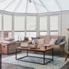 White cellular (honeycomb) blinds on tall bay windows and glazed roof, mostly lowered; filtering soft daylight; cozy conservatory living area with beige sofa, wooden coffee table, and blue-pattern rug.
