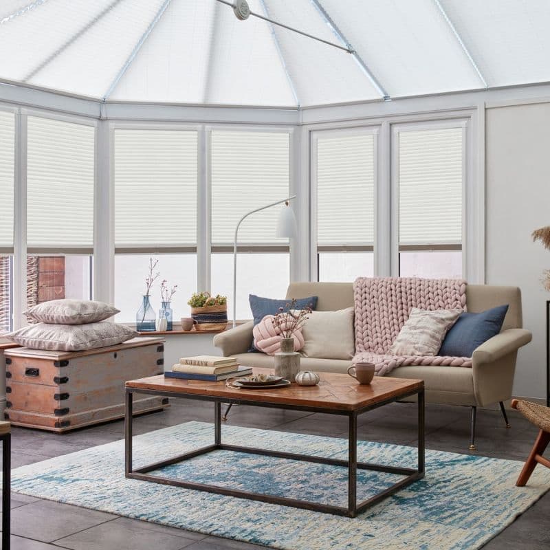 White cellular (honeycomb) blinds on tall bay windows and glazed roof, mostly lowered; filtering soft daylight; cozy conservatory living area with beige sofa, wooden coffee table, and blue-pattern rug.