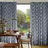 Navy-and-white leaf-patterned pleated curtains, parted to frame a door opening. They reveal sunlit backyard with trees and shed; foreground shows a wooden dining table set for breakfast under pendant lamp.