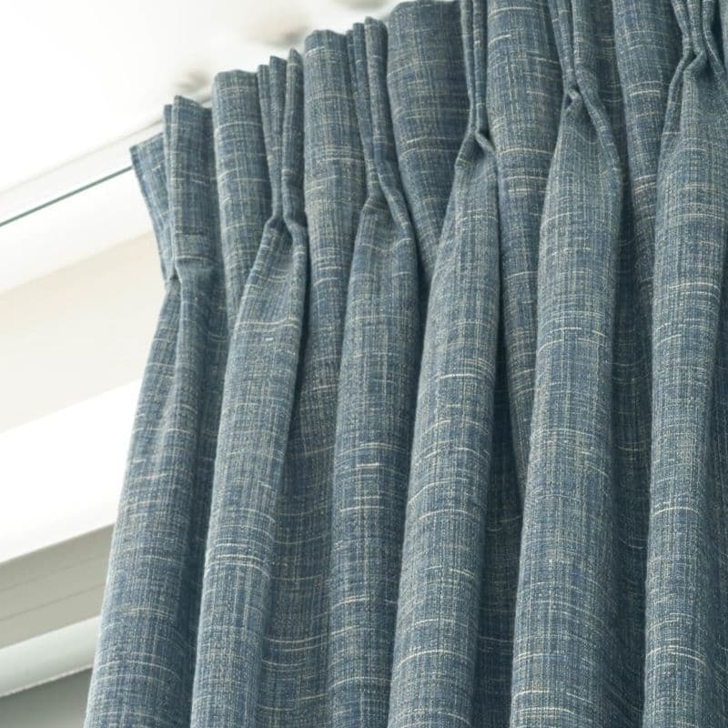 Blue, textured pinch-pleat curtains with white slub pattern, tightly gathered and hanging closed from a ceiling-mounted rod, filtering soft daylight inside a modern interior near a window.