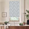 Blue patterned roller blind covers a window, filtering daylight above a stainless faucet and double sink in a modern kitchen with wooden cabinets, potted plants, and a framed sketch.