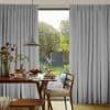 Grey pleated floor-to-ceiling curtains, parted at center in soft folds, filtering daylight; dining table with chairs and breakfast plates inside, backyard garden with trees and a swing visible outside.