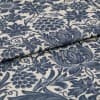 Blue floral-and-grape patterned upholstery fabric, folded to show thickness and weave, printed in indigo on a natural beige ground; close-up, studio-lit textile surface.