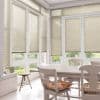 Beige horizontal Venetian blinds covering tall bay windows, lowered and filtering soft daylight; in a bright sunroom dining area with white table, chairs, floral cushions and garden view.