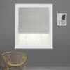 Grey Roman shade lowered with folded hem, partially filtering daylight; white-trimmed window set in a dark grey wall with a gold wire chair and small abstract framed art, blurred greenery.
