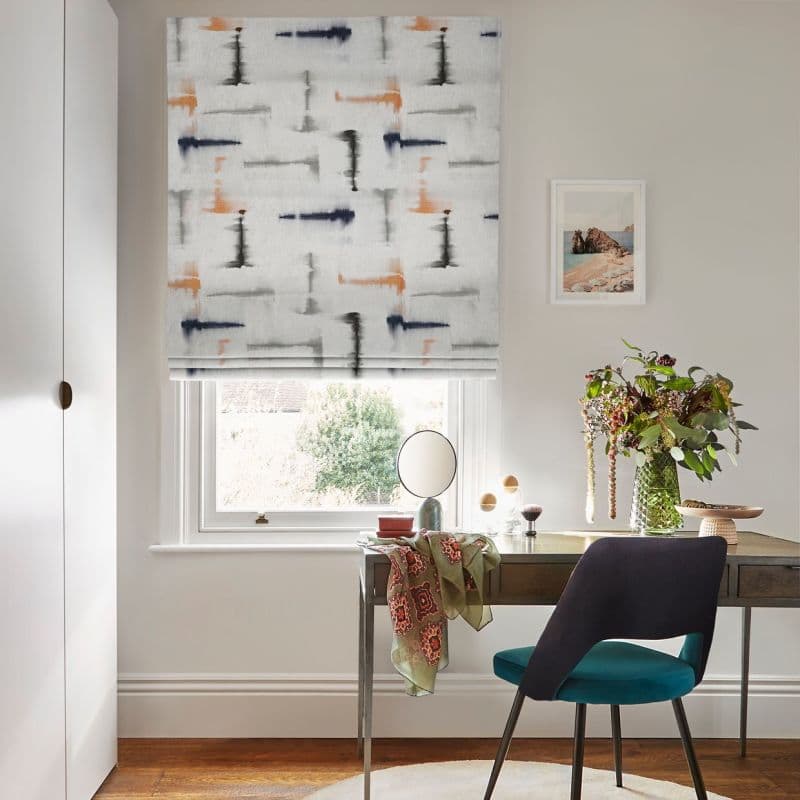 Roman blind—white fabric patterned with navy, gray and ochre brushstrokes—lowered, filtering soft daylight into a compact home office/vanity with desk, teal chair, vase of flowers and small garden view.
