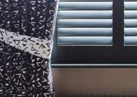 Black curtain with white triangular print, held back by a matching tieback; pulled aside to reveal closed horizontal shutters whose angled slats filter daylight; interior, windowsill visible.