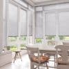 White horizontal blinds covering tall bay windows, lowered with thin slats partially closed, filtering soft daylight; pale dining table and chairs in bright breakfast nook facing a green garden.