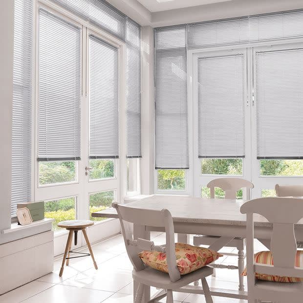 White horizontal blinds covering tall bay windows, lowered with thin slats partially closed, filtering soft daylight; pale dining table and chairs in bright breakfast nook facing a green garden.