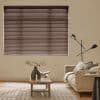 Brown horizontal Venetian blinds covering a large window, fully closed and filtering soft daylight through narrow slats; a neutral living room with beige sofa, floor lamp, wooden coffee table and rug.