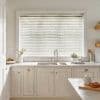 White horizontal Venetian blinds cover a large window, slats mostly closed and softly filtering daylight; above a farmhouse sink in a light, cream kitchen with marble counters, plants, and utensils.