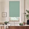 Teal roller blind covers most of a kitchen window, blocking light as sunlight filters below; stainless faucet, double sink, wooden cabinets, countertop with bowls, plants, framed sketch.