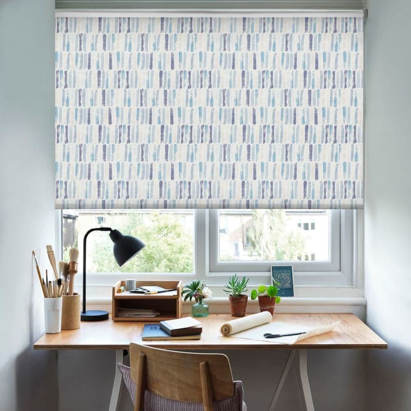 Patterned roller blind — white with vertical blue brushstroke motif — lowered across a double window, filtering soft daylight; a wooden desk beneath holds a lamp, potted plants, notebooks, and art tools.