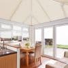 White pleated blinds cover the pitched conservatory roof, windows and French doors, mostly lowered and filtering daylight; small lower pane slightly raised, dining area with wooden table facing a garden.