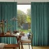 Teal pleated floor-to-ceiling curtains, drawn aside revealing central opening and soft filtered light; wooden dining table set with pancakes and plants beneath a pendant lamp, backyard garden and shed beyond.