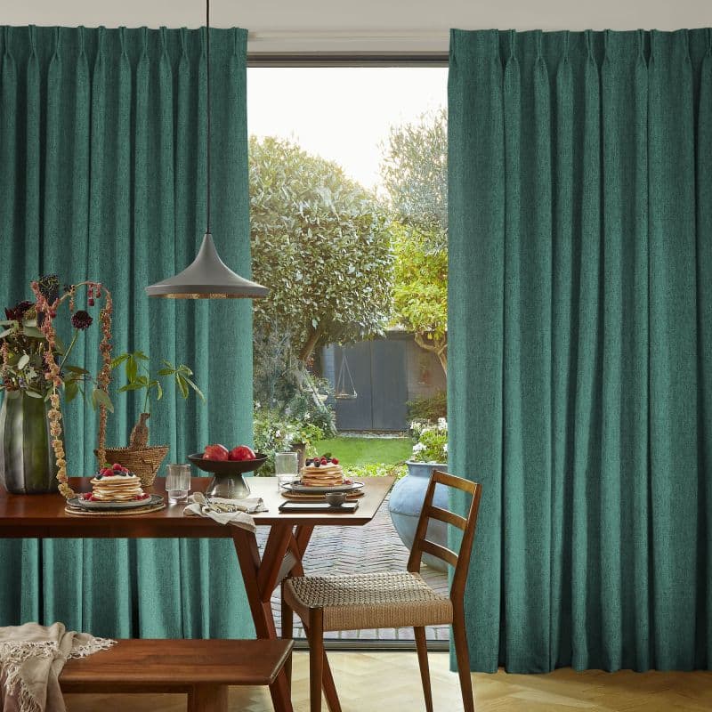 Teal pleated floor-to-ceiling curtains, drawn aside revealing central opening and soft filtered light; wooden dining table set with pancakes and plants beneath a pendant lamp, backyard garden and shed beyond.