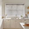 White horizontal venetian blinds covering a large window, slats mostly closed and slightly tilted to filter soft daylight; above a neutral, well-lit kitchen sink with marble countertop, chrome faucet, plants.