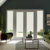 Pair of white French doors stand closed with lowered roller blinds, framing a sunlit garden beyond; living room features glass coffee table on rug, green velvet armchairs and wall art.